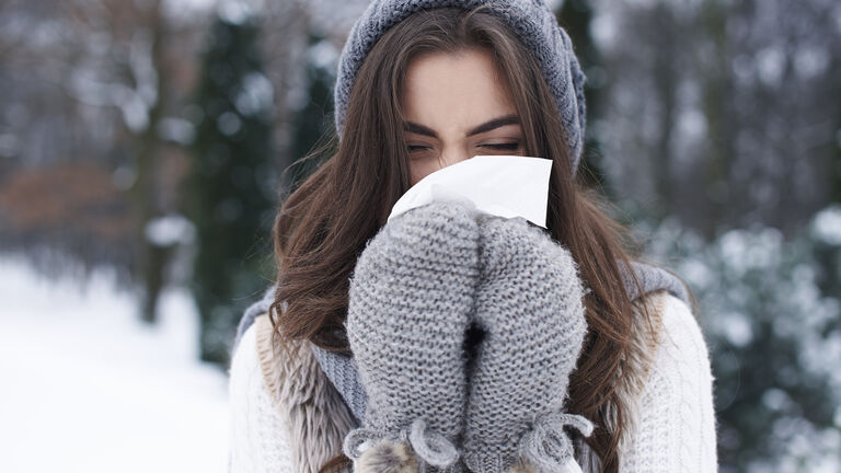Frau mit langen, braunen Haaren steht draußen im Schnee. Sie trägt Mütze und Fäustlinge und putzt sich mit einem Taschentuch die Nase