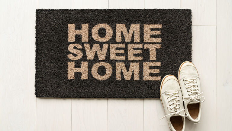 Schwarze Fußmatte für Eingangsbereich auf der in braucher Schrift steht "Home Sweet Home". Am rechen, unteren Fußmattenrand liegt ein Paar Damenturnschuhe.
