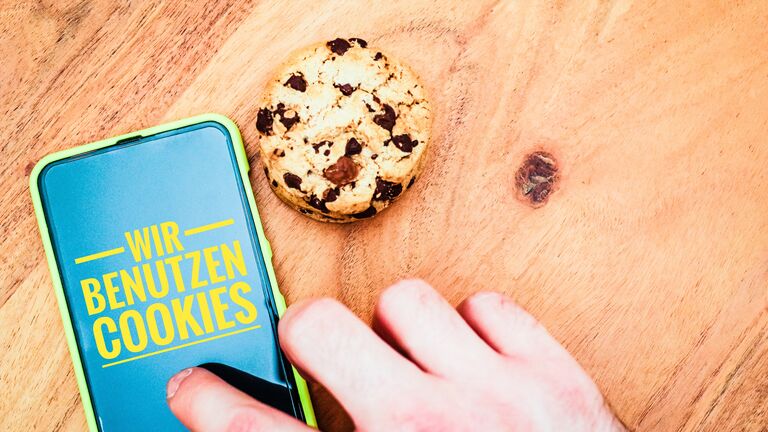 Eine Hand wischt über ein Smartphone auf dem "Wir benutzen Cookies" steht. Rechts neben dem Smartphone, welches auf einem Holzgrund liegt, ist ein Keks mit Schokoladenstücken platziert.