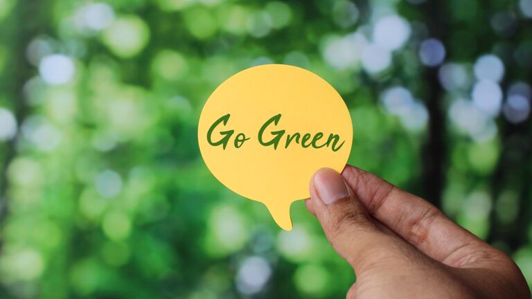 Hand hält einen Notizzettel in Sprechblasenform auf dem steht: Go Green (zu deutsch: Umweltfreundlich werden). Im Hintergrund sieht man Blätter und Bäume eines Waldes