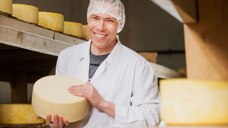 Mann mit weißem Kittel und Haube auf dem Kopf hält einen Laib Käse in der Hand. Im Hintergrund sind Holzregale mit weiteren Laiben Käse.