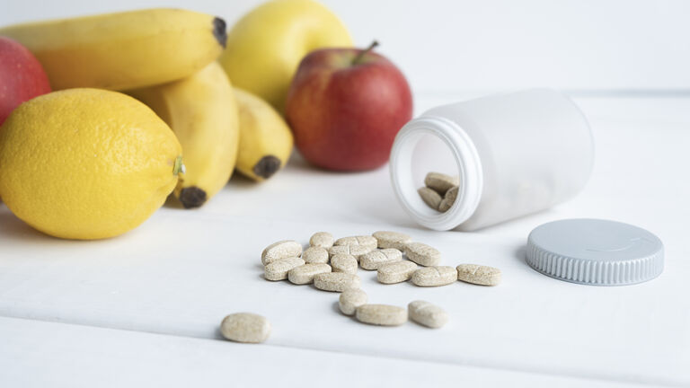 Eine Dose liegt geöffnet auf der Seite. Tabletten liegen auf dem Tisch. Im Hintergrund sieht man unscharf Bananen, Äpfel und Zitronen.