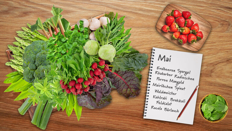 Saisonobst- und Gemüse wie Rhabarber, Spargel, Radieschen, Erdbeeren, Brokkoli, Mairübchen, Bärlauch, Kohlrabi, Porree, Feldsalat, Waldmeister. Spinat und weitere. Daneben ein Bleistift und ein Block, auf dem das Saisongemüse für Mai notiert ist. 