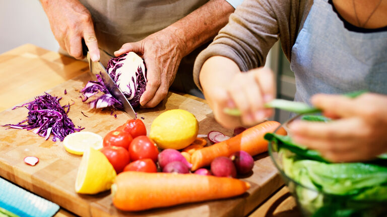 Zwei Personen schneiden Gemüse, wie Möhren, Rotkohl, Tomaten, auf einem Holzbrett klein