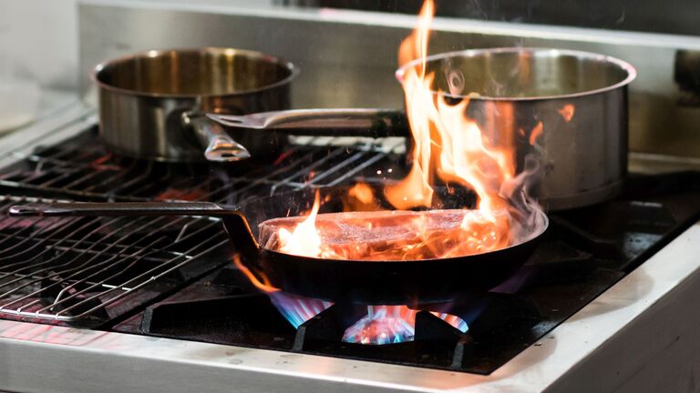 Aus einer Pfanne mit Fleisch auf eingeschaltetem Gasherd lodern Flammen