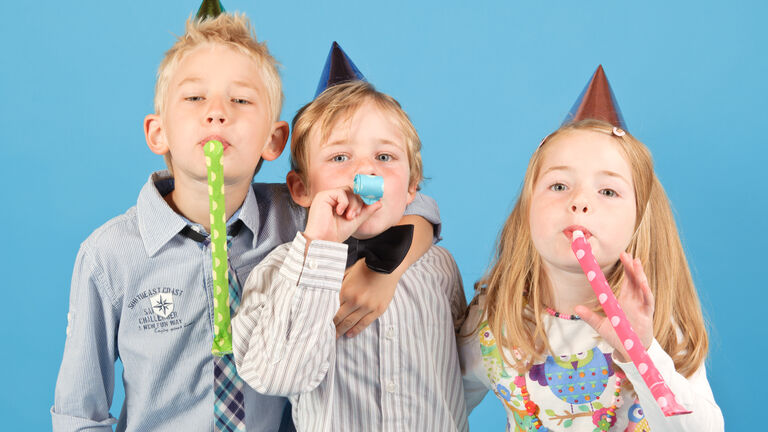 Drei Kinder, zwei Jungen und ein Mädchen, mit Partyhüten und Partytröten im Mund stehen neben einander.