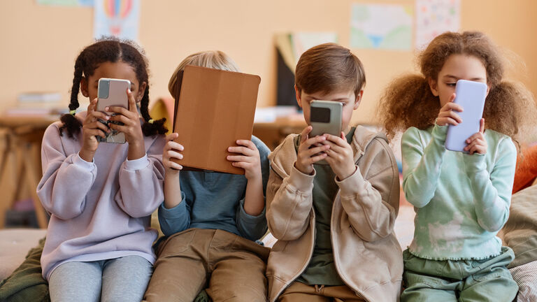 Vier Kinder im Alter von fünf bis sechs Jahren sitzen nebeneinander und halten entweder ein Smartphone oder eine Tablet vor das Gesicht.