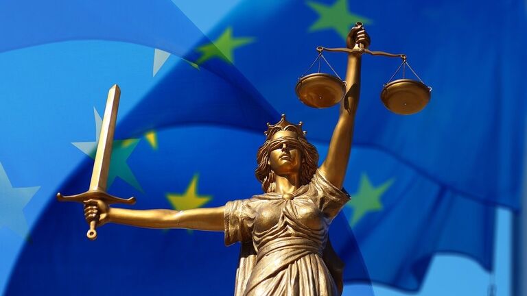 Die Justitia erhebt ihr Schwert und ihre Waage. Im Hintergrund weht die Flagge der Europäischen Union.