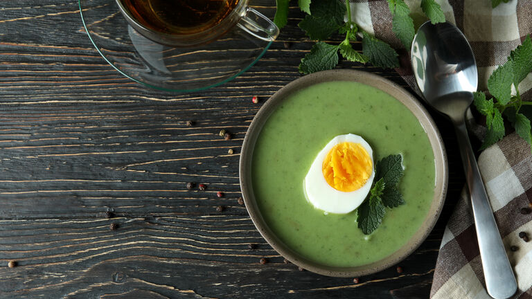 Brennnesselsuppe mit einem halben Ei garniert, von oben fotografiert. Daneben liegt ein Löffel. Weiter oben im Bild sieht man eine Glastasse mit Brennnesseltee.