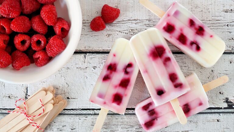 Selbstgemachtes Joghurt-Himbeereis aus einem Eisförmchen mit Holzstiel. Daneben steht eine Schüssel mit Himbeeren und ein Haufen Holzstiele.