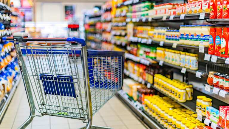 Ein Einkaufswagen steht zwischen zwei Supermarktregalen. In dem rechten Supermarktregal stehen Ketchup, Gewürzsoßen und Senf.