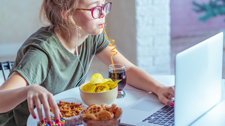 Eine junge Frau sitzt am Laptop und trink währenddessen mit einem Strohhalm aus einem Glas Cola. neben dem Glas Cola hat sie mehrere Schüsselchen mit Schokolinsen, Crackern und Salzstangen um sich herum stehen. Mit der rechten Hand greift sie während der Arbeit zu den Schokolinsen.