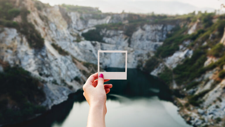 Ein Frauhand hält einen Polaroidbilderrahmen über eine Aussicht auf einen Fjord