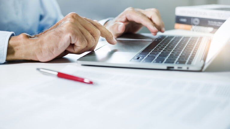 Eine Person sitzt am Schreibtisch vor einem Laptop. Man sieht nur Laptop und Hände die auf der Tastatur tippen. Im Vordergrund liegt ein Stift und ein Vertrag.