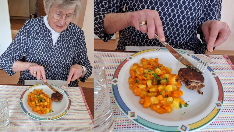 Seniorin sitzt am Essenstisch und ist Gemüse mit einer Frikadelle. Das Gericht ist vor ihr auf einem Teller angerichtet. Sie isst mit Messer und Gabel.