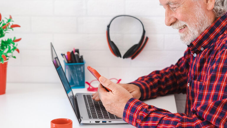 Ein älterer Herr sitzt an einem Tisch mit einem Laptop vor sich. Auf dem Tisch befindet sich zudem ein Stiftekorb. Der Herr bedient den Laptop und hat gleichzeitig ein Handy in der linken Hand.