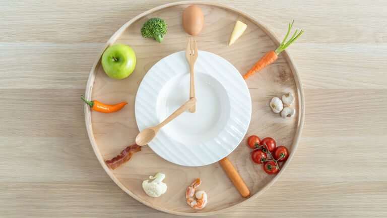 Ein Teller steht auf einem Tablett. Möhren, Tomaten, Knoblauch, liegen an den Stelle, wo bei einer Uhr die Zahlen sind. Das Gemüse liegt auf dem Tablett. Der Teller ist kleiner und auf ihm liegen Messer und Gabel als Zeiger. Sie zeigen 8 Uhr.