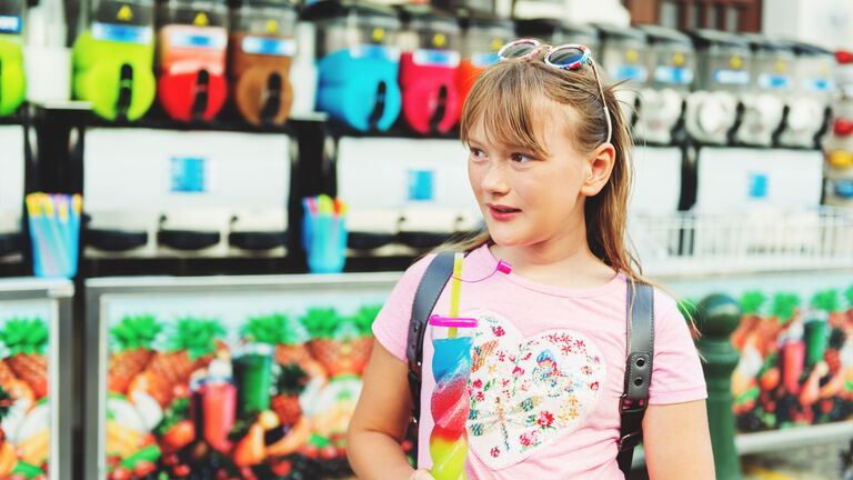 Ein kleines Mädchen steht mit einem Slush-Eis vor verschiedenenen Automaten mit unterschiedlichen bunten Slush-Eis-Sorten. Das Mädchen trägt einen Rucksack, ein T-Shirt und eine Sonnenbrille im Haar.
