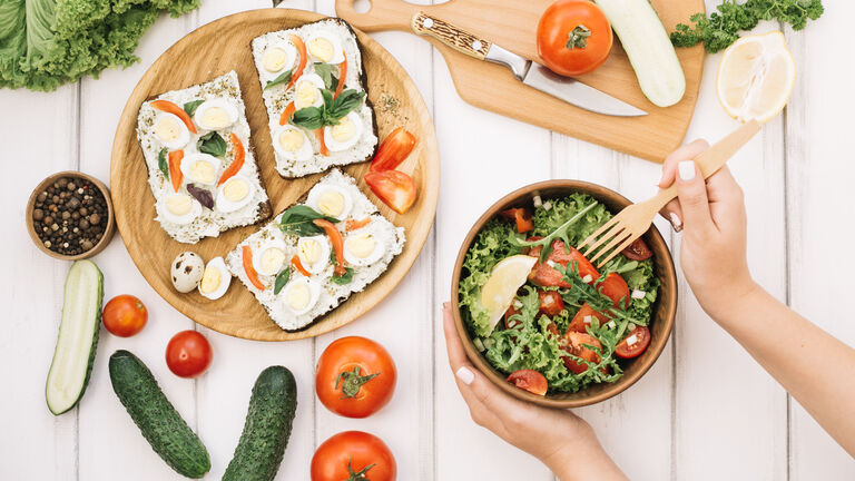 Auf einem Tisch sind unterschiedliche Speisen zu sehen. Belegte Brote, Tomaten, Gurken, Salat. In einer Schüssel ist aus den Zutaten ein Salat erstellt worden. eine Hand hält eine Schüssel und die zweite Hand piekst mit einer Gabel in die Schüssel.