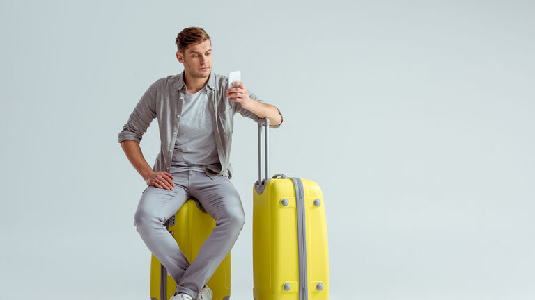 Ein jungern Mann sitzt auf einem gelben Koffer und schaut auf sein Smartphone in seiner linken Hand. Seinen linken Oberarm hat er auf den Griff eines weiteren gelben Koffer gestützt.