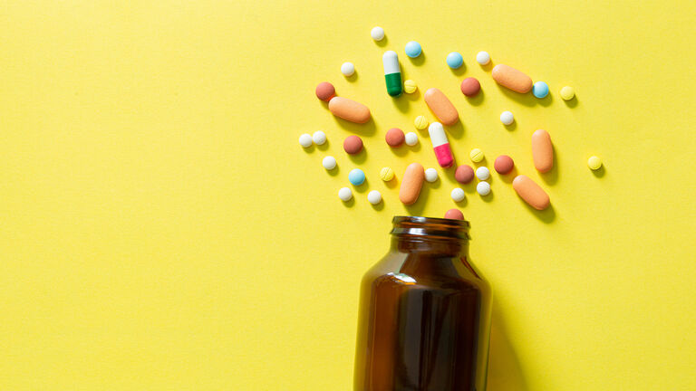 Eine braune kleine Glasflasche liegt auf der Seite auf einem gelben Untergrund. Mehrere runde, ovale und unterschiedlich bunte Tabletten liegen vor der Flaschenöffnung verteilt.
