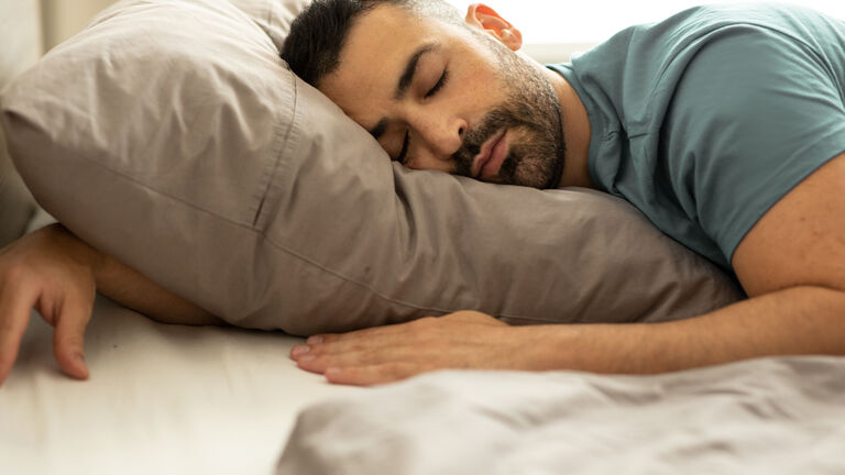 Ein Mann mit dunklen Haaren und Dreitagebart liegt auf dem Bauch im Bett. Eine Hand hat er unter dem Kopfkissen die andere Hand liegt direkt vor dem Kissen.