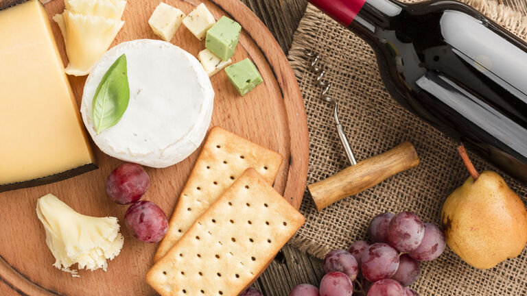 Auf einem Tisch steht ein Holzteller mit verschiedenen Schnitt- und Streichkäsesorten. Daneben liegen eine Flasche Rotwein, ein Korkenzieher und ein paar rote Trauben. Auf dem Teller liegen zudem noch Cracker.