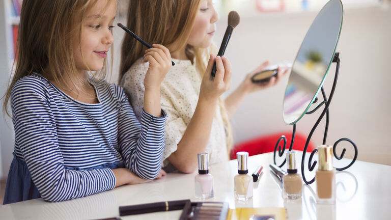 Zwei Mädchen sitzen vor einem Schminkspeigel und schminken sich. Ein Mädchen trägt Puder auf, das andere Lidschatten. Auf dem Tisch vor ihnen stehen mehrere Fäaschen Nagellack, Lidschatten und Mascara.