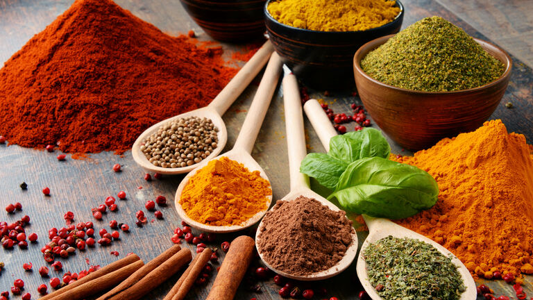 Verschiedene bunte Gewürze auf weißen Löffeln. einige Gewürze sind auch in braunen und weißen Schälchen im Hintergrund. Außerdem gibt es zwei Gewürzhaufen link und rechts im Bild.