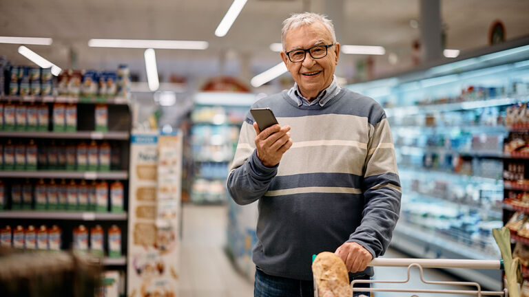 Ein älterer Herr steht mit Einkaufswagen im Supermarkt und hält in der rechten Hand ein Smartphone. Der Herr schaut fröhlich in die Kamera.