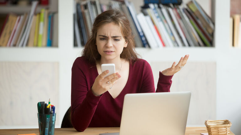 Eine junge Frau sitzt vor einem Bücherregal vor ihrem Rechner und hat ein Smartphone in der Hand. Ihr gesichtsausdruck ist verärgert und frustriert.