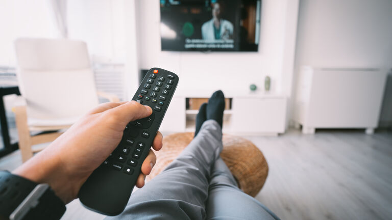 Aus der Sicht eines Mannes auf einem Sofa: Er hat die Beine auf einen Schemel gekreuzt gelegt und hat in der linekn Hand eine Fernbedienung. Im Hintergrund sieht man einen Fernseher an der Wand. Die Szene ist in einem Wohnzimmer.