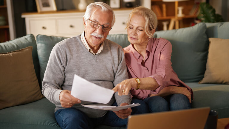 Ein älteres Paar sitz konzentriert lesend auf einer Couch und schaut in Unterlagen. Die Frau zeigt auf eine Stelle in den Unterlagen.