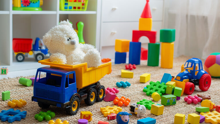 Ein Blick in ein Kinderzimmer: Auf dem Boden liegen viele bunte Plastikbauklötze sowie ein Plastik-Trecker und ein Plastik-LKW indem ein Plüschteddy sitzt. Im Hintergrund sieht man einen Turm, der aus Plastikbauklötzen gebaut wurde. Außerdem kann man im Hintergrund ein Regal sehen, in dem ebenfalls Plastikspielzeug, wie beispielsweise ein Plastik-LKW und ein Plastikkorb mit Bällen drin steht. Das regal ist weiß.