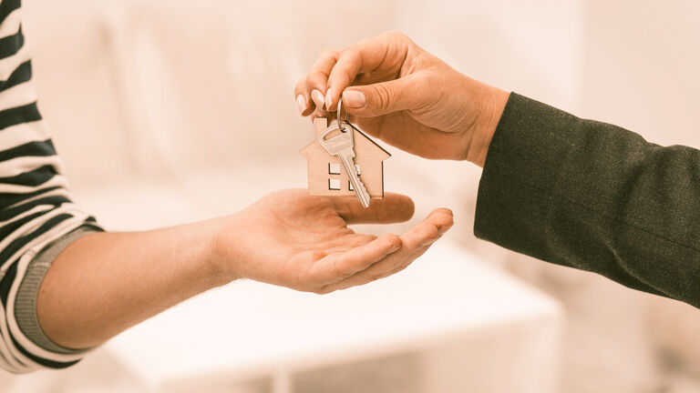Zwei Hände: Die eine Hand gibt der anderen Hand einen Schlüssel mit Schlüsselanhänger. Der Schlüsselanhänger ist aus Holz und hat die Form eines Häuschens.