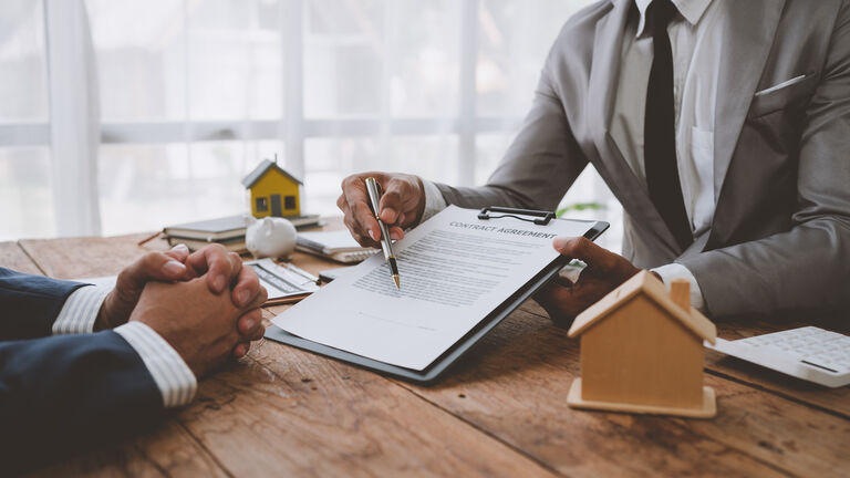Ein Blick auf einen Schreibtisch. Rechts zeigt ein Mann in einem grauen Anzug mit einem Kugelschreiber auf einen Vertrag auf einem Klemmbrett. Ihm gegenüber sitzt eine weitere Person im Anzug - man sieht allerdings nur die Hände. Auf dem Schreibtisch stehen zwei Häuschen aus Holz.
