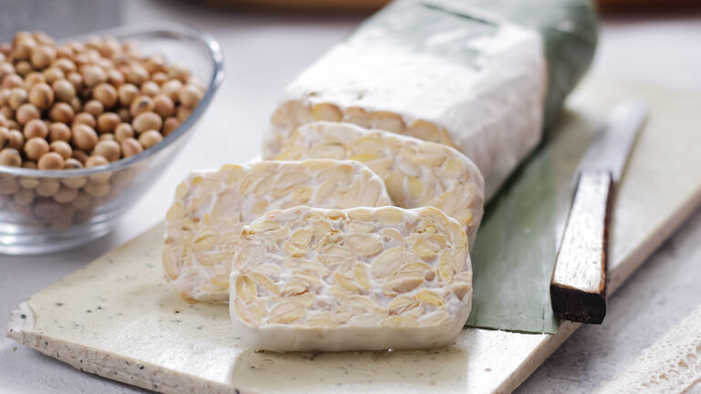 Ein Tempehblock liegt auf einem Marmorbrett. Drei Scheiben sind bereits abgeschnitten und fächern sich vor dem Block auf. Neben dem Tempeh liegt ein Messer. Rechts davon steht eine Glasschale mit Sojabohnen drin.