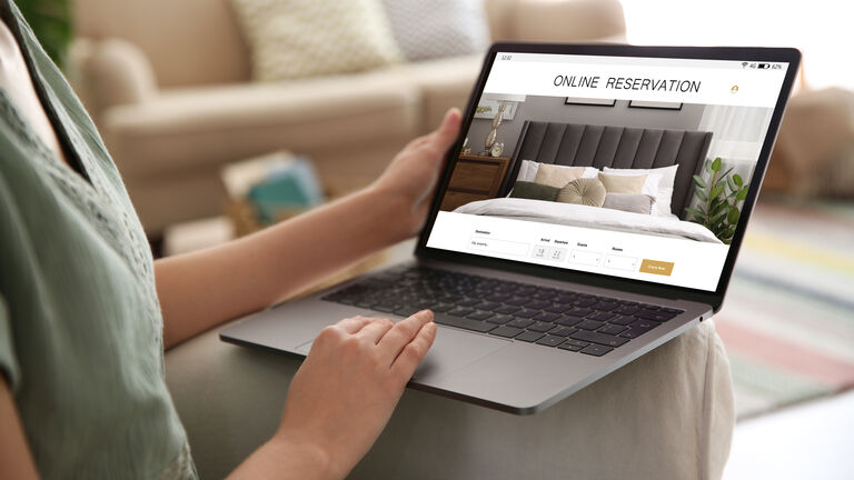 Eine Frau sitzt im Wohnzimmer und hat ihren Laptop auf dem Schoß. Sie hat eine Webseite geöffnet, auf der man Hotelreservierungen vornehmen kann.