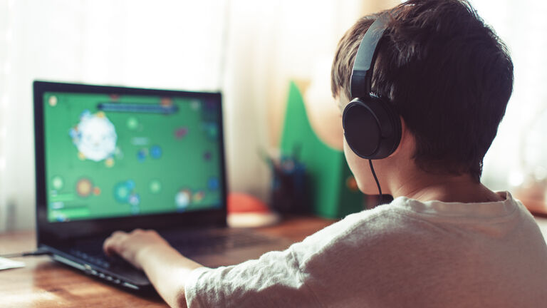 Ein Kind mit kurzen Haaren und Kopfhörern von hinten, dass am Laptop ein Computerspiel spielt.