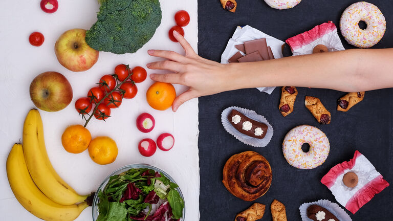 Zweigeteiltes Bild: Links sind gesunde Lebensmittel wie Salat, Cocktailtomaten, Bananen, Äpfel, Mandarinen, Bananen und Radieschen zu sehen; rechts hingegen Kuchen, Donuts, Schokolade und anderes Gebäck. Eine Hand greift von rechts ins Bild und greift sich eine Mandarine von der linken Seite.