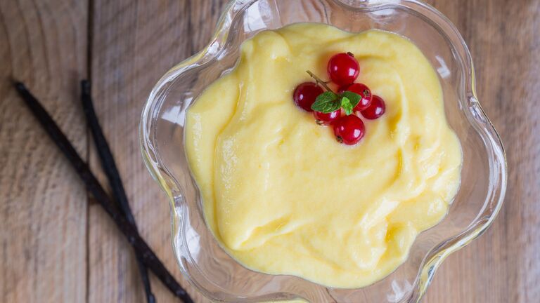 Glasschälchen mit Vanillepudding und Johannisbeere als Deko, daneben zwei Vanilleschoten