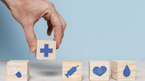 Es liegen nebeneinander aufgereiht einzelne Holzwürfel mit Symbolen rund um das Thema Medizin. Eine Hand hält einen weiteren Würfel in der Hand.