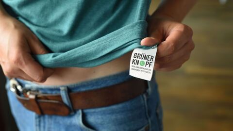 Ein Mensch hält sein T-Shirt so, dass das Label Grüner Knopf zu sehen ist