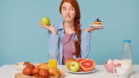 Frau mit verschiedenen Lebensmitteln Frau sitzt am Tisch, vor ihr liegen verschiedene Lebensmittel von Milch, Organensaft und Mufins über Obst und Gemüse. In ihrer rechten Hand hält sie einen Apfel. In ihrer linken Hand hält sie ein Törtchen. Sie macht einen kritischen Gesichtsausdruck.