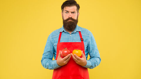 Ein bärtiger Mann in Küchenschürze hält Paprika und Tomaten in der Hand