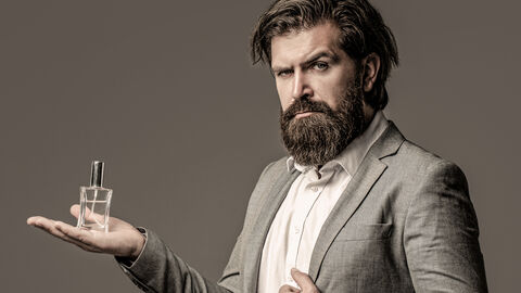 Ein bärtiger Mann im Anzug hält eine Flasche Parfum in der flachen Hand