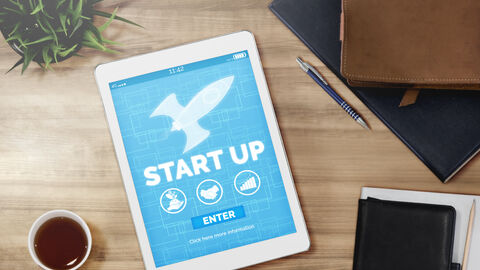 Auf einem Tisch liegt ein Tablet Computer mit der Aufschrift Start-Up und wird von Kalender und Tasse umringt