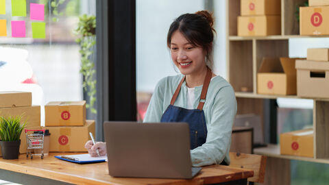 Eine junge Frau verpackt Artikel in einen Versandkarton. Im Hintergrund ist ein Regal mit weiteren Kartons
