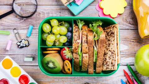 Brotdose mit Schulbrot, Gemüse, Krackern und Obst auf einem Tisch mit Stiften, Malkasten, Lupe und Notizzetteln