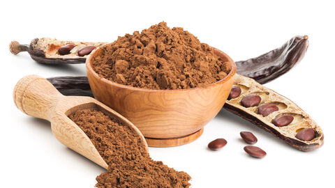 Carobschote neben Carobpulver in einer Holzschale und einem Holzmesslöffel